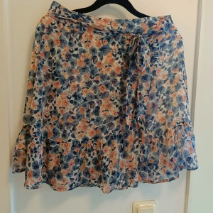 Blommig omlottkjol med volang - Supersöt kort omlottkjol med blommigt mönster i blått, lila och persikofärgat. Kjolen har volangkant och knyt i midjan för en extra snygg touch. Perfekt för dig som gillar romantisk och lekfull stil.