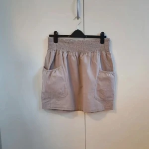 Beige kortkjol från Ellos Fresh Spirit - Säljer en beige kortkjol från Ellos Fresh Spirit med bred smock i midjan och två stora fickor framtill. Kjolen är gjord i mjuk bomull och har en avslappnad passform som är både chill och snygg. Perfekt för dig som gillar enkel och clean stil.