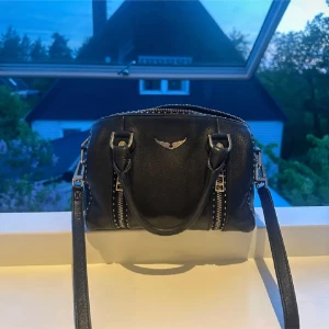 Zadig&Voltaire väska - Säljer en svart handväska från zadig voltaire med silverfärgad logga och dragkedjor framtill. Väskan har både handtag och axelrem, vilket gör den enkel att bära på olika sätt. Tillverkad i skinn med snygga detaljer och stilren design.