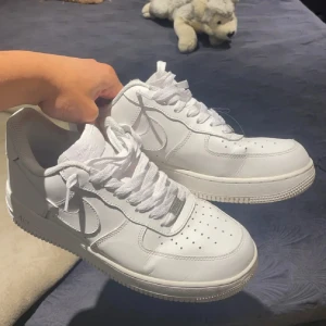 Nike Air Force 1 helvita sneakers - Klassiska Nike Air Force 1 sneakers i helvitt skinn. Snygg och clean design med perforerad tå, rund tå och platt sula. Ikonisk Nike-logga på sidan och AIR-detalj på hälen. Perfekta för dig som vill ha en tidlös och fräsch look.