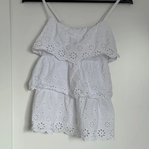 Vit kort topp från Robin - Supersöt vit topp från Robin med smala axelband och broderade blommor i spets. Klänningen har volangdetaljer och är tillverkad i mjuk bomull, perfekt för varma dagar. Den är kort och har en luftig passform som ger en chill vibe.