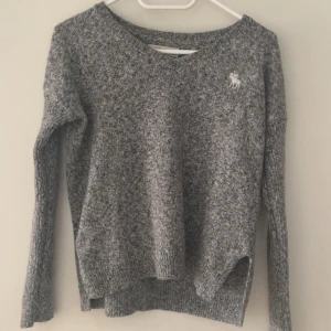 Grå stickad tröja Abercrombie - Mysig gråmelerad stickad tröja från Abercrombie med v-ringad hals och lång ärm. Tröjan har en liten vit logga broderad på bröstet och ribbade muddar vid ärmslut och nederkant. Perfekt för chill dagar och när du vill vara extra comfy.
