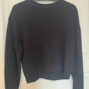 Svart stickad tröja med grov struktur - En svart stickad tröja med grov ribbad struktur och rund hals. Tröjan har en loose passform och långa ärmar, perfekt för lager-på-lager under kyliga dagar. Enkel och stilren design som funkar till många olika outfits.