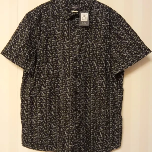 Svart blommig kortärmad skjorta Dressmann - Kortärmad skjorta från Dressmann i svart med små blommiga mönster i vitt och beige. Skjortan är tillverkad i 100% bomull och har klassisk krage och knäppning framtill. Perfekt för dig som gillar en stilren men ändå lekfull look.                                              Kan skickas med extra pris via post.