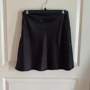 Svart satin kjol från Chiquelle - Snygg svart kortkjol från Chiquelle i glansigt satinmaterial. Kjolen har en enkel och stilren design med lätt A-linjeformad siluett. Perfekt för dig som gillar minimalistisk stil och vill ha en kjol som känns både trendig och bekväm.