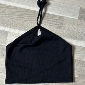 Svart halterneck topp från Zara - Snygg svart halterneck topp från Zara med ribbad struktur och knyt i nacken. Toppen har en liten droppformad öppning framtill som ger en cool detalj. Perfekt att styla med jeans eller kjol för en trendig look.