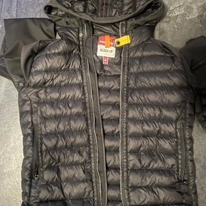 Svart pufferjacka Parajumpers M - Svart pufferjacka från Parajumpers med huva och coola detaljer som gul rem vid halsen. Jackan har dragkedja framtill och två sidofickor med dragkedja. Perfekt för dig som vill ha en stilren och sportig look.