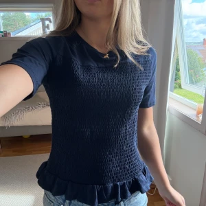 Mörkblå smockad blus med volang - Supersöt mörkblå blus med smockat tyg över hela framsidan och en volangkant längst ner. T-shirtrmar och rund halsringning ger en chill vibe. Perfekt till jeans eller kjol för en snygg look.