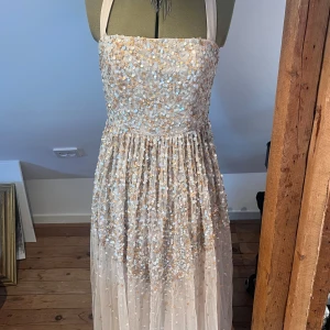 Champagnefärgad långklänning med paljetter - Magisk långklänning i champagnefärg med glittrande paljetter i guld och silver. Klänningen har breda axelband som knyts i nacken och ett skirt, flowy tyg som ger en drömmig känsla. Midjebandet är löst så man kan antingen ta bort den eller sy på den igen! Storlek M men skulle säga passar S bättre. 