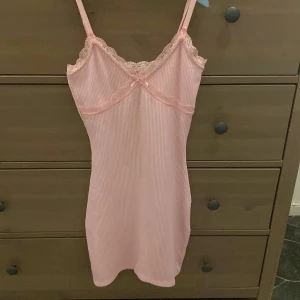 Rosa ribbad kortklänning med spets - Supersöt ljusrosa kortklänning med smala axelband och spetsdetaljer vid urringningen. Klänningen är ribbad och har en liten rosett framtill, vilket ger en gullig och feminin vibe. Perfekt för dig som gillar en enkel men söt stil.