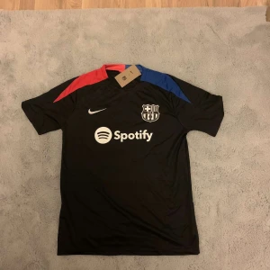 FC Barcelona svart Nike fotbollströja - Säljer en svart FC Barcelona fotbollströja från Nike med vita loggor och sponsortryck från Spotify. Tröjan har korta ärmar och detaljer i rött och blått på axlarna. Materialet är lätt och andas, perfekt för träning eller match. Klassisk Barca-sköld på bröstet.