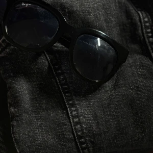 Svarta solglasögon från Haga Eyewear - Snygga svarta solglasögon från Haga Eyewear med bred båge och klassisk form. Glasögonen har en stilren look med mörka linser och passar perfekt till en trendig streetstyle. Ramen är i blank svart plast och ger en cool vibe.