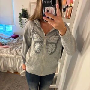 Grå hoodie med fjäril och strass - Grå hoodie med dragkedja och huva, dekorerad med en stor fjäril i strass på framsidan. Tröjan har snörning i huvan och praktiska fickor framtill. Perfekt för dig som gillar lite extra bling.