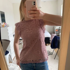 Rosa spetsblus med volangärmar - Säljer en söt rosa blus i spets med volangdetaljer vid ärmarna och hög hals. Blusen är ärmlös och har ett romantiskt mönster i spetsmaterial. Perfekt att styla med jeans eller kjol!