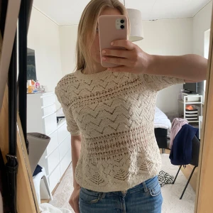 Beige stickad topp med spetsdetaljer - Säljer en beige stickad topp med korta ärmar och spetsmönster. Toppen har ett luftigt, virkat utseende med fina detaljer och rund hals. Perfekt att styla med jeans eller kjol för en trendig look.