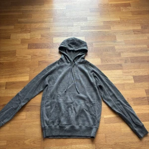 Grå/brun hoodie med dragsko - En enkel och stilren grå hoodie med huva och dragsko. Tröjan har långa ärmar och en klassisk magficka framtill. Perfekt för en avslappnad look.