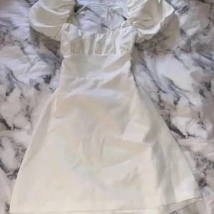 Vit kort puffärmad studentklänning med öppen rygg - Superfin vit klänning med puffärmar och öppen rygg med snörning. Klänningen har en söt fyrkantig urringning och är kort i modellen. Perfekt för studenten, endast använt en gång