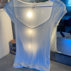 Vit topp med spetskant från Nelly - Säljer en tunn och söt vit topp från Nelly i storlek XXS. Toppen har korta ärmar, v-ringning och en fin spetskant runt halsen. Perfekt för varma dagar eller som layering-plagg.