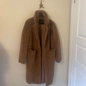 Lång teddyjacka från Vero Moda - Mysig lång brun teddyjacka med djupa fickor i storlek S. Den är rätt oversized så passar även M. Endast provad, aldrig använd utomhus så i nyskick. Köpt för ca 6-700kr på Vero Moda men säljer nu för 100kr då den ej används. Jackan har en rak passform och stängs med knappar framtill. 