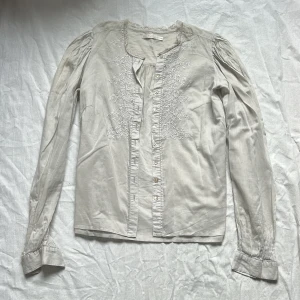 Beige blus med broderi från Wera - Säljer en beige blus från Wera med broderade blommor framtill. Blusen har långa ärmar med lätt puff vid axlarna och knappar längs hela framsidan. Rundad halsringning med spetsdetalj. Storlek 38 men liten i storleken. 
