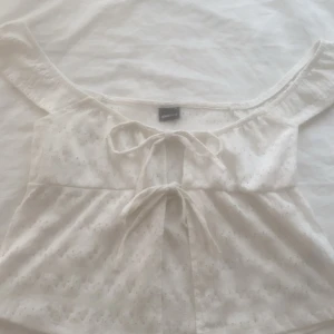 Vit offshoulder topp med knyt - Söt vit topp från Gina Tricot med offshoulder-modell och två knyt framtill. Toppen har ett diskret hålmönster i tyget och är perfekt för varma dagar. Tillverkad i bomull och har en luftig och feminin känsla.