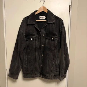Svart jeansjacka från NA-KD - Säljer en svart jeansjacka från NA-KD i klassisk modell med knappar framtill och två bröstfickor med lock. Jackan har långa ärmar och en avslappnad passform, perfekt att slänga över en hoodie eller t-shirt.