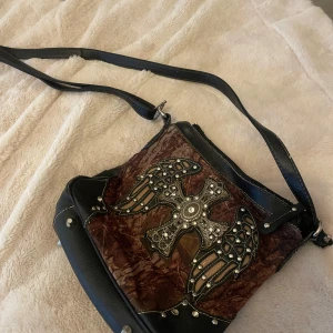 Brun och svart axelväska med kors och nitar - Cool axelväska i brun sammet och svart syntet med stort kors och vingar dekorerat med nitar och stenar på framsidan. Väskan har justerbar axelrem och dragkedja.  Lite sliten på baksidan av väskan syns på sista bilden 💗