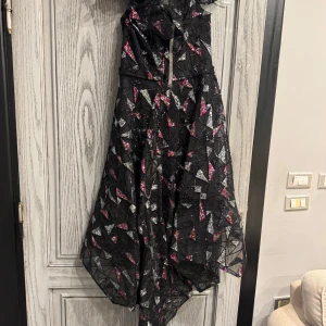 Svart glittrig långklänning med asymmetrisk fåll - Unik svart långklänning med glittriga silver- och rosa detaljer i geometriskt mönster. Klänningen har en asymmetrisk fåll, dekorativ blomdetalj vid axeln och ett stort glittrigt spänne i midjan. Perfekt för dig som vill sticka ut.