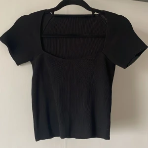 Svart ribbad topp med kort ärm - Säljer en svart ribbad topp med korta ärmar och fyrkantig ringning. Toppen har en figurnära passform och är tillverkad i ett stretchigt material som sitter snyggt på kroppen. Perfekt att matcha med jeans eller kjol.