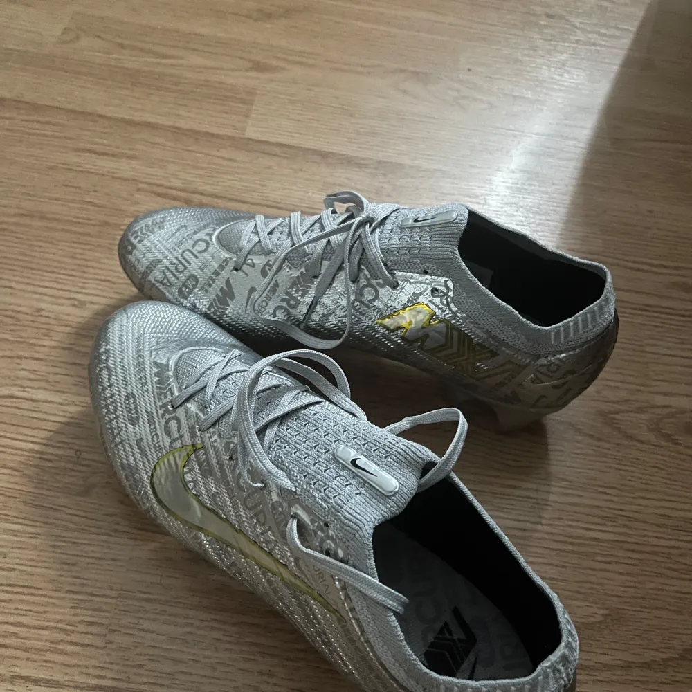 Säljer ett par Nike Mercurial fotbollsskor i metallic silver med guldigt märke på sidan. Skorna har snörning och mönstrad ovandel med textdetaljer. Yttersulan är i blankt silver med fasta dobbar, perfekt för spel på gräs och konstgräs!. Kengät.