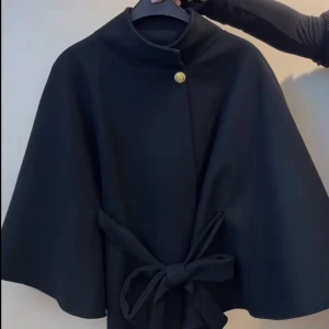 Svart poncho med knytning och guldknapp - Stilren svart poncho från Missäy med cape-form, hög krage och dekorativ guldknapp vid halsen. Som man även kan vika ner. Ponchon har ett  knytbälte i midjan som ger en snygg siluett. Perfekt för dig som vill ha en elegant och tidlös look. Har använts endast få gånger 💕