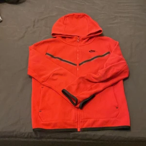 Röd hoodie från Nike med dragkedja - Säljer en röd hoodie från Nike med hel dragkedja och svarta detaljer. Jackan har huva, långa ärmar och en praktisk ficka med dragkedja på ena ärmen. Perfekt för en sportig look.