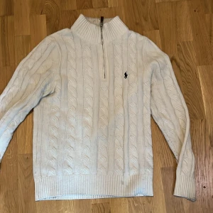 Polo Raulph Lauren Stickad Tröja - Säljer en vit kabelstickad tröja från Polo Ralph Lauren med halv dragkedja och hög krage. Tröjan har storlek M men passar som S. Perfekt för lager-på-lager under kyliga dagar.