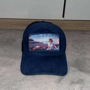 Mörkblå Monaco keps från Revenite - Snygg mörkblå keps med mesh-bak och tryck framtill som föreställer en kvinna och en sportbil. Justerbar passform och märket Revenite syns på insidan. Perfekt för dig som vill sticka ut med en unik keps. Kepsen är limiterad och tillverkas inte mer.