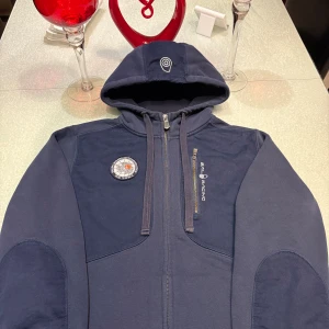 Blå Sail racing  hoodie med dragkedja - Mörkblå hoodie från Sail Racing med hel dragkedja och huva. Jackan har en broderad logotyp på bröstet och en rund patch. Mjukt fleecefoder på insidan och förstärkta partier på armbågarna.