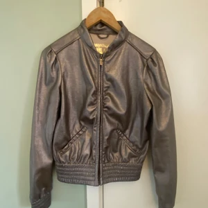 Metallicgrå bomberjacka - Säljer en metallicgrå bomberjacka med dragkedja framtill och resår i midja och ärmslut. Jackan har två snedställda fickor med knapp och en cool glansig finish. För liten på mig på sista bilden 