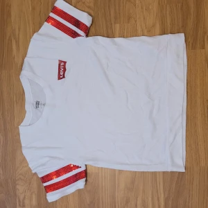 Vit t-shirt med röda paljettärmar från Levi's - Vit t-shirt från Levi's med korta ärmar dekorerade med röda paljetter och vita ränder. Klassisk passform och liten röd Levi's-logga på bröstet. Perfekt för dig som vill sticka ut med lite extra glitter i vardagen.