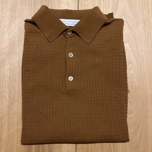 Brun pikétröja från FILIPPO DE LURENTIIS - Säljer en brun pikétröja med klassisk krage och tre knappar framtill. Tröjan har korta ärmar och är tillverkad i ett strukturerat bomullstyg som ger en snygg och unik känsla.