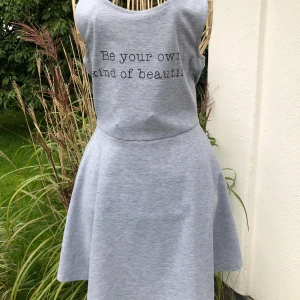 Grå ärmlös klänning med text - Säljer en ljusgrå ärmlös klänning med rund halsringning och texten 'Be your own kind of beautiful' framtill. Klänningen har en utställd kjol och är tillverkad i mjukt bomullsmaterial. Perfekt för varma dagar!stl 36 från Divided