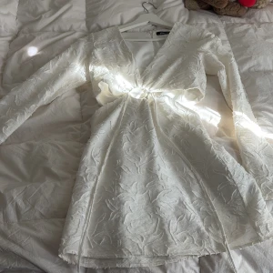 Vit kortklänning från Gina Tricot - Supersnygg vit kortklänning från Gina Tricot med lång ärm och strukturmönster. Klänningen har v-ringning, markerad midja med knytband och en öppen detalj framtill. Perfekt för sommarens tillfällen!