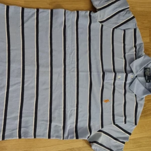 Randig pikétröja från Polo Ralph Lauren - Snygg ljusblå pikétröja med mörkblå och vita ränder från Polo Ralph Lauren. Klassisk krage, korta ärmar och broderad logga i orange på bröstet. Perfekt för en avslappnad och stilren look.