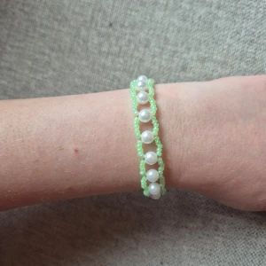 Handgjort armband med pärlor och gröna pärlor - Säljer ett unikt armband med vita pärlor i mitten och små ljusgröna pärlor runtom. Armbandet har ett flätat mönster och är perfekt som accessoar för att ge din outfit en söt touch.