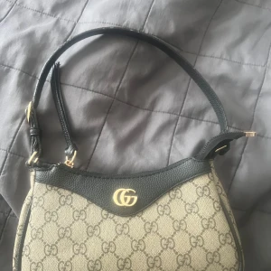Beige och svart handväska  - Säljer en snygg beige och svart handväska med klassiskt GG-mönster och guldfärgad logga framtill. Väskan har svarta detaljer och axelrem i skinn. Perfekt accessoar för att lyfta din outfit. 