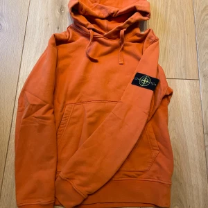 Orange hoodie från Stone Island - Säljer en orange hoodie från Stone Island med klassisk patch på ärmen. Tröjan har huva med dragsko och en stor magficka framtill. Perfekt för dig som gillar streetwear och vill sticka ut med färg.