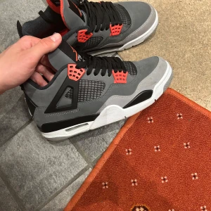 Nike Air Jordans 4 Retro i grå och svart - Säljer ett par Nike Air Jordans 4 Retro i grå mocka med svarta och röda detaljer. Skorna har klassisk snörning, meshpaneler på sidorna och vit sula med synlig Air-enhet. Perfekta för dig som gillar streetwear och sneakers med ikonisk design.
