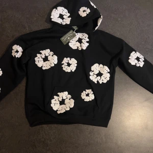 Denim Tears hoodie - Säljer en svart hoodie från Denim Tears med stora vita blommor tryckta över hela tröjan. Tröjan har huva och en klassisk passform, perfekt för dig som vill sticka ut med en unik design.