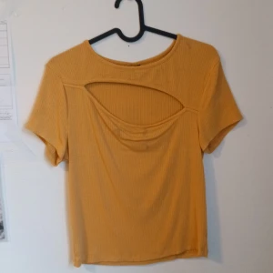 Gul ribbad topp med cut out från Gina Tricot - Säljer en gul/orange ribbad topp från Gina Tricot med korta ärmar och snygg cut out-detalj framtill. Toppen har rund halsringning och är i ett mjukt, stretchigt material. Perfekt för att addera lite färg till din outfit!