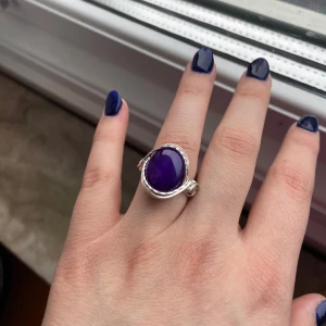 Silverring med lila sten - Säljer en unik silverring med en stor, oval lila sten i mitten. Ringen har en snygg, virad design runt stenen som ger ett coolt och trendigt intryck. Perfekt statement piece för dig som vill sticka ut!