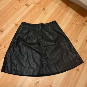 Svart kjol i skinnimitation från Vila - Säljer en svart kjol i skinnimitation från Vila. Kjolen har en lätt A-linjeform, resår i midjan och dragkedja bak. Perfekt för dig som gillar en stilren look med lite edge.