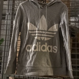 Grå hoodie från Adidas - Säljer en grå hoodie från Adidas med stor vit logga framtill. Tröjan har långa ärmar, huva med dragsko och är tillverkad i mjukt material. Perfekt för en avslappnad och sportig stil. Lite nopprig 